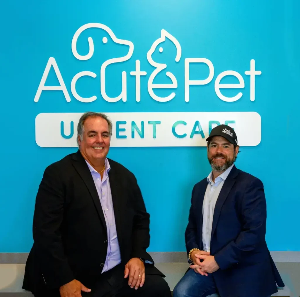 AcutePet Founders, Drs. Doug Hoffman & David Sachs sitting on a counter in front of the AcutePet Urgent Care Logo on a teal wall